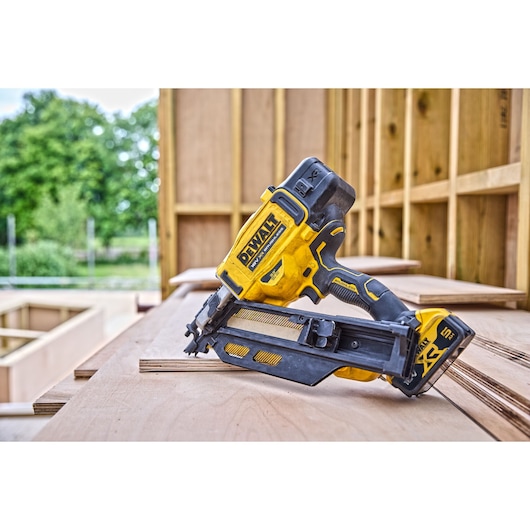 DCN930 18V XR Brushless framing nailer stands on top of a wooden surface in an open construction site. Trees are visible in the background