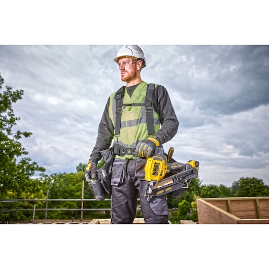 Construction worker standing on top of a rooftop. A DCN930 18V XR Brushless framing nailer is suspended from his toolbelt